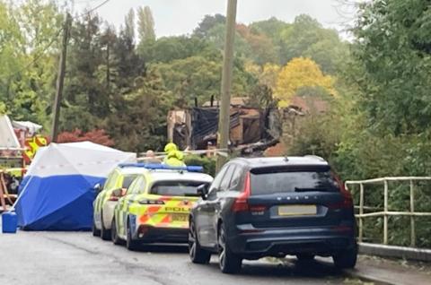 Two police cars, a parked navy-coloured car, a forensic tent and a fire engine beside what in the distance appears to be a partially collapsed brick house with collapsed roofing.