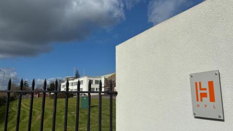 A crooked black metal fence is in the foreground, with a large expanse of grass behind it, leading to a single storey factory building on the right of the picture. Behind that is another series of building with cars parked in front. On the right of the picture in the foreground is a white wall with the HPL Prototypes logo on it 