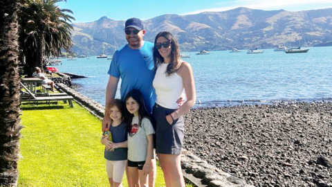 A man, a woman and two young girls standing next to a lake with mountains in the background. It is sunny - the adults are wearing sunsglasses.