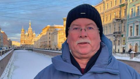 Headshot of David Crisp taken in St Petersburg. He has a grey moustache and rimless glasses and is wearing a black woollen beanie hat, a blue puffer coat, with a black jumper and other layers underneath. He is standing on a bridge over a frozen canal with an ornate church in the distance and buildings lining the canal. The sky is blue with soft sunlight on some of the buildings. 