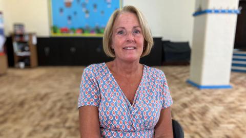 Heather Curtis wears a pink, red and blue dress is sitting on a chair and smiling at the camera. She is in the school. The background is blurred. 