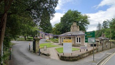 The entrance to a hospice site. There is a yellow and white branded signage, and a footpath leading to a single-storey building. The entrance is set back from the road, which has double-yellow lines running along it.