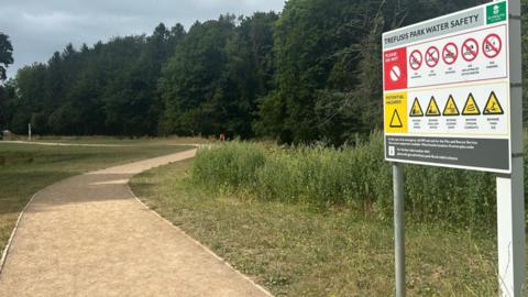 A picture taken from the entrance to Trefusis Park. There is a big sign advising water safety and a large path carved around grass with large green trees in the background.
