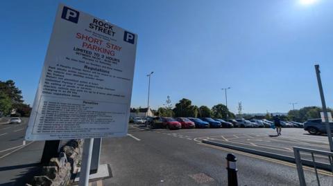 Cars parked in a car park. The sky is blue. The cars are on the right, on the left is a wall saying Rock Street short stay parking.