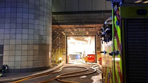 A fire engine parked inside a large industrial building, with hoses laid across the floor leading towards a brightly lit open doorway, scaffolding around the entrance, and tiled concrete pillars on either side.