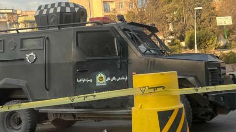 An armoured vehicle is parked in front of yellow tape and a yellow plastic street blockade.