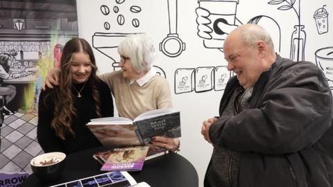 An older man and woman sit at a cafe table with a younger girl. The woman has her arm around the girl and she is holding a comic book the pair are looking at. The man is also looking in their direction and they are all smiling. There are more comic books on the table, next to a hot drink. In the background on the wall there are comic book style drawings.