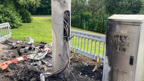 A mass of scorched rubble and plastic lays at the bottom of a 5G tower. To the right of the tower is a grey electricity box which is also scorched black. The words 'open your eyes' have been painted on the electricity box. A mass of loose electricity wires are hanging from an opening in the mast. 