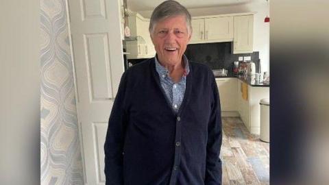 John Hardy is standing in front of a kitchen smiling. He has grey hair and is wearing a blue shirt and long cardigan.