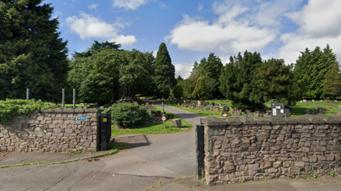 The entrance to a cemetery, there is a stone wall between the entrance. Then green grass with trees and a path through the middle. There are headstones on either side of the path. The sky is blue and cloudy in the background. 