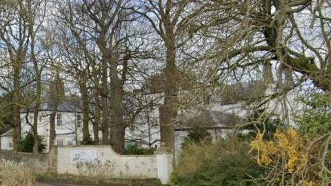 Howlish Hall care home is a white building tucked behind a row of trees and a white fence. 