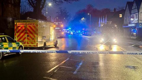 Police tape blocks off a road at night with emergency vehicles seen behind the cordon. 