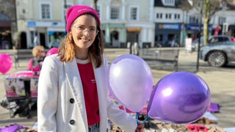 A woman holding two purple balloons is stood in a high street. She is wearing a pink t-shirt and a white jacket with glasses and a pink beanie hat.