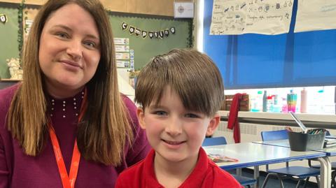 A mother and her young son smile at the camera as they sit in a school classroom. She has long brown hair and is wearing a purple pullover, while he has short brown hair and is wearing a red polo shirt. 