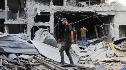A man wearing a black t-shirt and trousers walks through the rubble of a destroyed building in Gaza City.