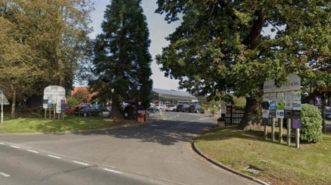 An entrance into a garden centre. There are tall green trees either side of the road and two signs in front that read "Bell plantation".