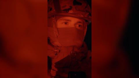 The face of a man wearing a balaclava and an army helmet, who is lit up in red light at night.