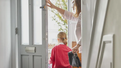 A young girl is walking out of her front door with her mother behind her. She has a red jumper on and her mother has a stripey top on. Furniture in the hall area is white/cream.