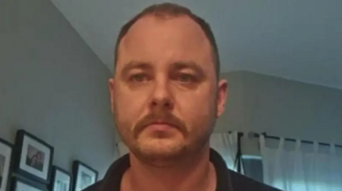 A man looks into the camera in close-up. He is in a home, we can see some pictures hanging on the wall behind him and a set of curtains. He has very short cropped hair and a moustache. 