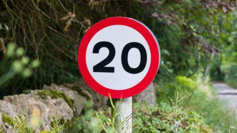 A 20mph road sign on the left of in an Oxfordshire lane, which is free of traffic or anyone walking down it.
