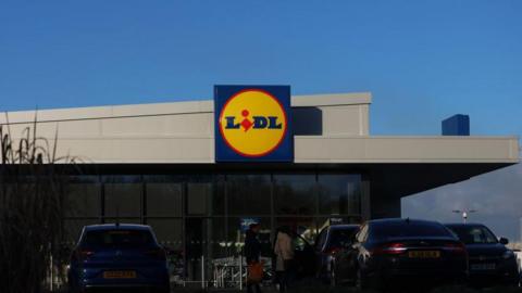 A generic photo of a Lidl supermarket, the building is grey, there are cars in a car park parked out the front. In the middle is the blue, yellow and red Lidl logo. 