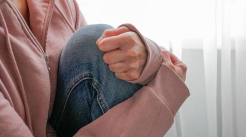 A woman is wearing a pink fleece and blue jeans. Her head is not visible. White curtains are in the background.