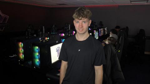 A teenage boy stands in the centre of the frame staring directly at the camera. He is wearing a black shirt and has blonde hair. Behind him are rows of gaming computers, glowing with a range of coloured lights. There are students in chairs playing games on these computers.