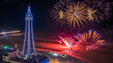 Aerial view of the fireworks display on the beach beside Blackpool's North pier with crowds of people watching on the promenade in the evening with the Blackpool Illuminations on and Blackpool Tower lit up purple in the background to the left.