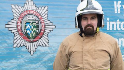Fire station manager Tom Wright stands in front of a side of a fire engine. He is wearing beige firefighting uniform with a white helmet. The side of the panelled engine is blue and has a red and green Cumbria Fire and Rescue Service badge.