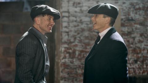 Two actors in formal dress wearing flat caps in conversation. One man is Cillian Murphy stood on the right, the other is Barry Keoghan.