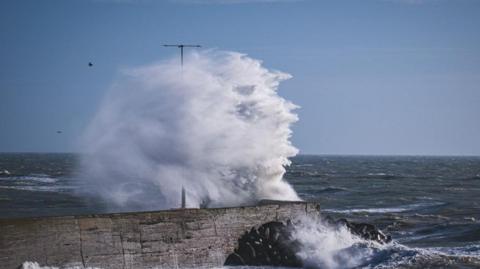 Waves crashing high over harbour walls. The sea is visible in the background