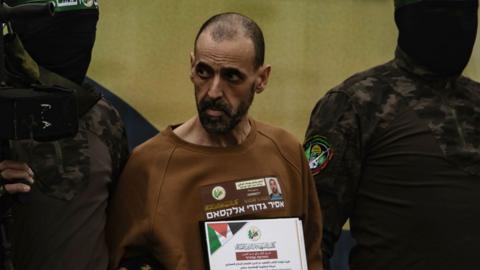 Eli Sharabi is in the middle, being held by two hooded men in black and khaki clothing. He is gaunt and wearing a brown sweatshirt. He has a facial hair in the shape of a goatee and a balding hair line. He is holding a sign which has Arabic writing and a Palestinian flag in the corner