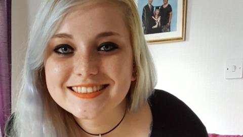 A smiling Rebekah Morris with dyed blonde hair in front of a picture from her graduation ceremony