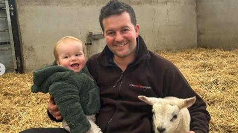 A man sits on a barn floor holding a young boy in one arm and a young sheep in another. 