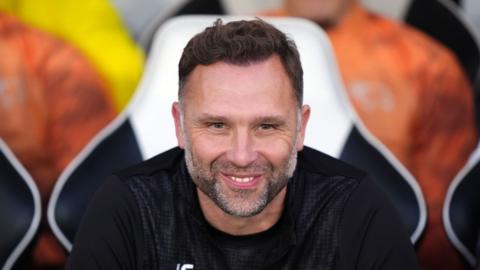 Derby County head coach John Eustace looks ahead smiling wearing a black training top.