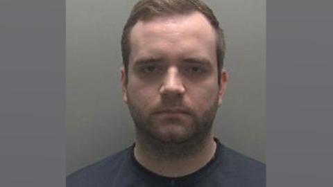 A slightly blurry police mug shot of Thomas Verdicchio, who has short light brown hair and stubble. He is wearing a dark blue T-shirt. 