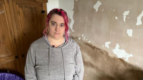 A woman with pink hair and a grey hoodie stands in front of a mouldy wall, without paint