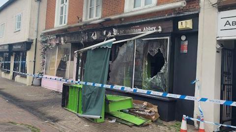 A burnt shop front, with broken windows, with police tape along the front. The signs to the shop are fire damaged, and items are outside on the pavement, as well as a green awning that has come down. 