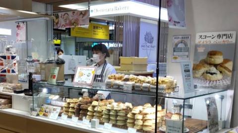 Under the bright lights of a railway station in Japan, there are lots of scones stacked up with prices and descriptions in Japanese and a server waiting 
