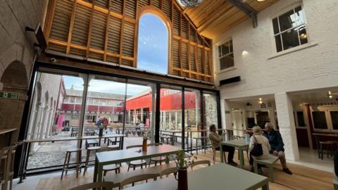 A market containing a seating area for the public and a courtyard is. People are sat at the picnic like benches in the middle of the market. There are large windows.