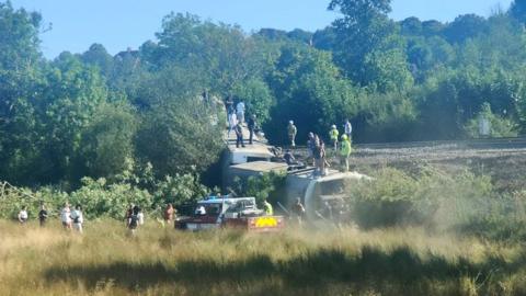 A derailed train with people stood on top, surrounded by a field.
