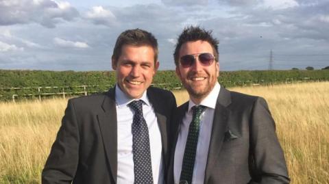 Jonathan and George Rudd wearing dark suits, light shirts and ties, smiling towards the camera, standing in front of a field of hay grass with a long hedgerow behind them