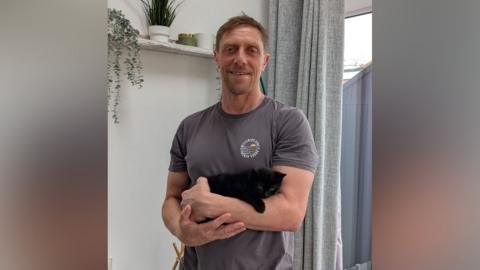 A man with a grey T-shirt holding a tiny black kitten in his arms. He is standing inside a house with a curtain in the background.