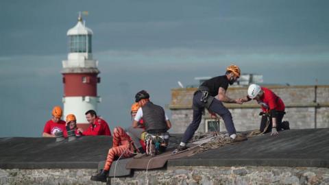 Two air ambulance supporters dressed in red t-shirts sit on the Citadel wall while two abseil instructors dressed in dark clothing assist them.