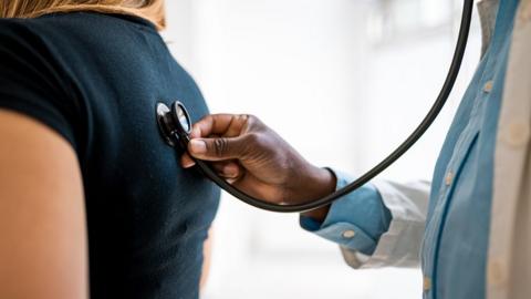 A doctor holds a stethoscope to the back of a female patient