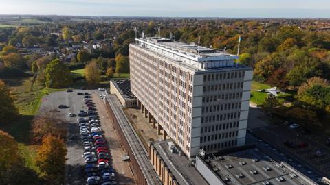 A drone image of a large office building in a rectangular shape, with trees, a car park and housing around it.