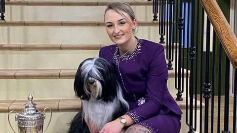 Paige Le Moignan and Mika sit on a set of tiled stairs with black metal banisters either side of them. Paige is wearing a purple-above knee length dress with diamantes covering the neckline and the hem. Mika is black and white in colour and has hair covering her eyes. They are sat next to their trophy which is silver with large handles and is about 20 inches tall. Next to the trophy is a red and gold rosette.