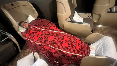 A man tries out a fully reclining pod on a ferry, he has dark hair and is covered in a red and black patterned blanket