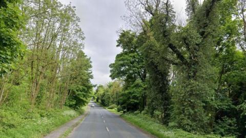 A rural road with just one car on it, in the far distance. Trees flank the road on both sides.