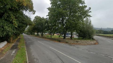 A semi-rural tree lined stretch of road, with a junction in the foreground and car park visible through trees